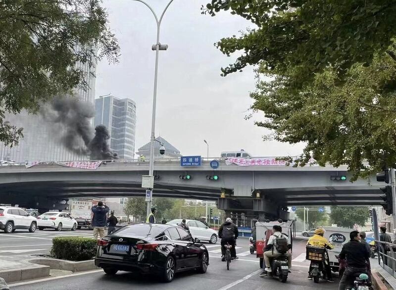 People watch while smoke rises as a banner with a protest message hangs off Sitong Bridge, Beijing, China October 13, 2022 in this image obtained by Reuters.